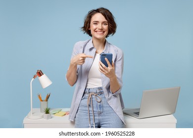 Young happy fun secretary employee business woman 20s in casual shirt work stand at white office desk with pc laptop point index finger on mobile cell phone isolated on pastel blue background studio.