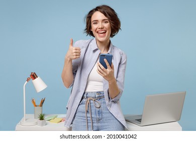 Junge fröhliche Sekretärin Geschäftsfrau Geschäftsfrau in der Freizeitshirt-Arbeit steht an weißem Schreibtisch mit PC Laptop halten Handy-Show Daumen-up Geste einzeln auf pastellblauem Hintergrund Studio.