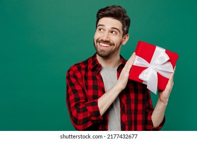Young happy fun smiling man he 20s in red shirt grey t-shirt hold red present box with gift ribbon bow look aside on workspace isolated on plain dark green background studio. People lifestyle concept - Shutterstock ID 2177432677