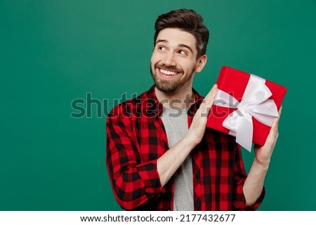 Young happy fun smiling man he 20s in red shirt grey t-shirt hold red present box with gift ribbon bow look aside on workspace isolated on plain dark green background studio. People lifestyle concept