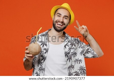 Young happy fun tourist man in beach shirt hat hold cocktail juice in coconut bowl with straw show v-sign isolated on plain orange background studio portrait. Summer vacation sea rest sun tan concept