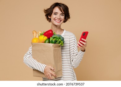 Young Happy Fun Vegetarian Woman 20s In Casual Clothes Hold Paper Bag With Vegetables Hold In Hand Use Mobile Cell Phone Read Browsing Menu Isolated On Plain Pastel Beige Background Shopping Concept