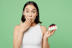 Young Happy Fun Woman Wear White Clothes Holding In Hand Pice Of Cake Dessert Cover Mouth Isolated On Plain Pastel Light Green Background. Proper Nutrition Healthy Fast Food Unhealthy Choice Concept