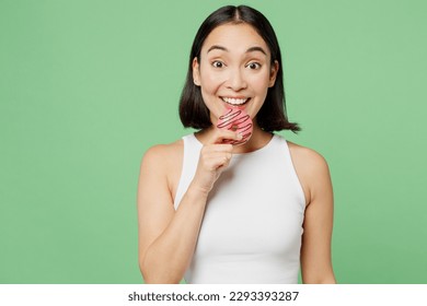Young happy fun woman wearing white clothes hold in hand biting donut dessert look camera isolated on plain pastel light green background. Proper nutrition healthy fast food unhealthy choice concept
