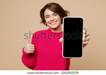 Young happy fun woman wearing pink sweater hold use close up mobile cell phone with blank screen workspace area show thumb up isolated on plain pastel beige background studio. People lifestyle concept