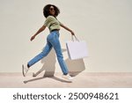 Young happy funky African American woman customer shopper in sunglasses jumping walking on white background holding shopping bag buying retail fashion clothes making online sale purchase. Full body.