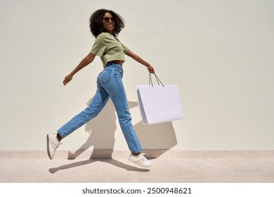 Young happy funky African American woman customer shopper in sunglasses jumping walking on white background holding shopping bag buying retail fashion clothes making online sale purchase. Full body. - Powered by Shutterstock