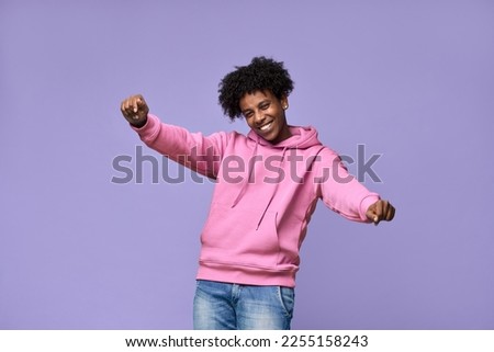Young happy funky African American teen guy wearing pink hoodie having fun isolated on light purple background. Smiling cool ethnic generation z teenager student model dancing and moving.