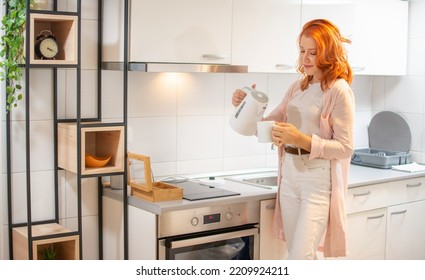 Young Happy Ginger Woman Making Tea With Electric Kettle In The Kitchen At Home.