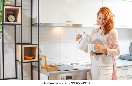 Young happy ginger woman preparing tea with electric kettle in the kitchen at home. - Powered by Shutterstock