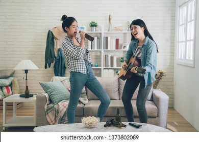 Young happy girl best friends having party one playing guitar and another singing by holding beer bottle as microphone. two asian women dancing together. Friendship leisure rest home enjoy concept