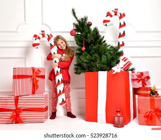 Young happy girl in Christmas Santa hat with big present gift smiling near huge red box and holding big candy cane on a white background. New year 2017 concept