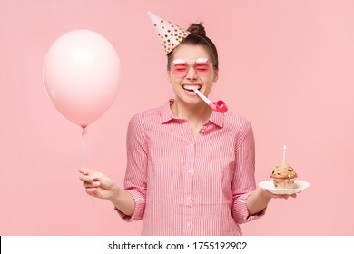 Young happy girl in colored glasses and birthday hat, blowing whistle, holding pink balloon and cupcake, laughing and enjoying party, isolated on pink background - Powered by Shutterstock