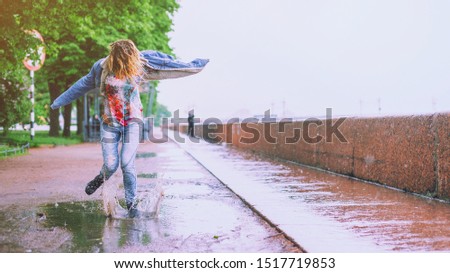 Young happy girl jumping in a puddle on the road under summer rain. Positive funny woman splashing water legs on a rainy day in the city. Feet in shoes or rubber boots. Spring season and rain concept