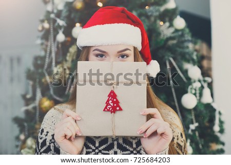 Young happy girl in red santa hat covering half face with christmas gift on xmas tree background. Funny and joyful winter holiday wallpaper
