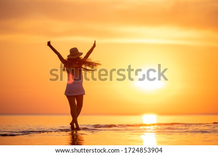 Young happy girl is standing or dancing at sunset beach with raised arms