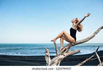 Young  happy girl in swimsuit sitting on old tree snag rise hands high in air, posing on black sand sea beach. Travel lifestyle concept. Retreat leisure on summer family vacation in tropical island - Powered by Shutterstock