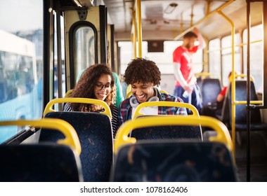 Young Happy Gorgeous Couple Is Sitting Together In A Bus And Smiling While Looking At Something Hidden Behind The Bus Seat.