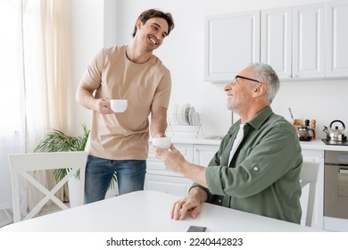 young and happy guy giving coffee cup to smiling grey haired father sitting in kitchen - Powered by Shutterstock