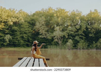A young happy hippie woman sitting on the lake shore on a bridge in eco clothing made of natural materials in harmony with nature in the fall