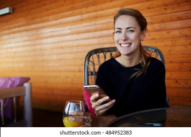 Young Happy Hipster Girl With Mobile Phone In Hands Is Looking To The Camera, While Is Sitting In Modern Coffee Shop Interior.Cheerful Female Student Is Using Cell Telephone During Rest After Lectures