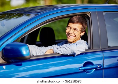 Young happy hispanic man wearing glasses and blue jeans shirt sitting behind wheel of his car and smiling through window - new drivers concept