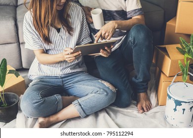 Young Happy Homeowner Couple Moving Into New House, Sitting And Relaxing On The Floor And Looking For Home Decorating Ideas And Shop Online On Tablet