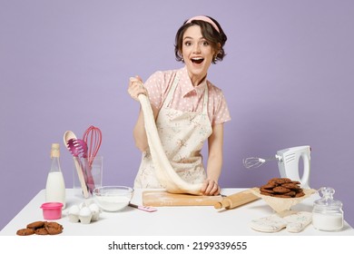 Young Happy Housewife Housekeeper Cook Chef Baker Woman In Pink Apron Work At Table Kitchenware Kneads Dough Baking Isolated On Violet Background Studio Portrait Process Cooking Food Pastry Concept.