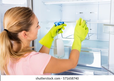 Young Happy Housewife Woman Cleaning The Empty Refrigerator Door With Spray Bottle And Sponge