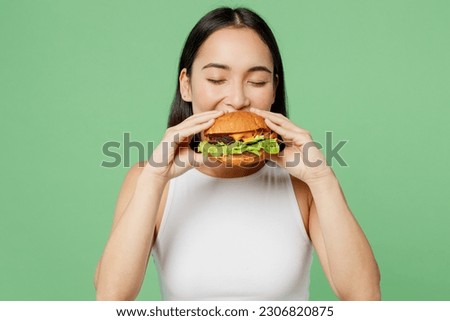Young happy hungry fun cheerful woman wear white clothes holding eating biting tasty burger isolated on plain pastel light green background. Proper nutrition healthy fast food unhealthy choice concept