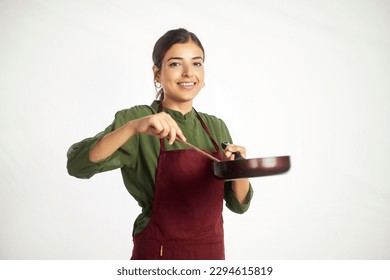 A young happy Indian Asian modern woman wearing an apron is holding a wooden spatula and preparing cuisine in a frying pan looking at the camera and smiling. Cooking, kitchen, homemade food concept 