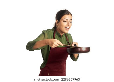 A young happy Indian Asian modern woman wearing an apron is holding a wooden spatula and preparing cuisine in a frying pan looking at the camera and smiling. Cooking, kitchen, homemade food concept  - Powered by Shutterstock