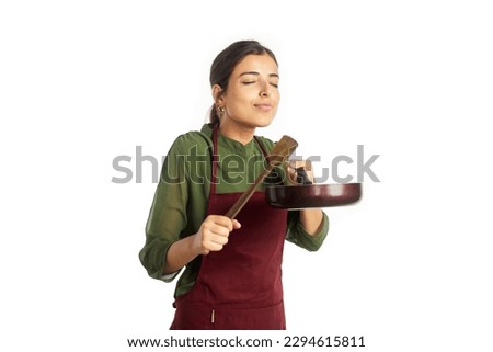 A young happy Indian Asian modern woman wearing an apron holding a wooden spatula and frying pan is smelling the aroma of freshly prepared cuisine. Concept of cooking, kitchen, homemade food