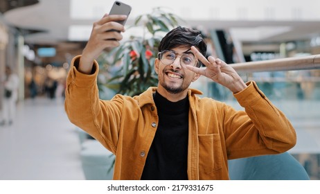 Young happy indian guy blogger holds smartphone recording vlog filming video for blog in mall joyful man waving greeting in webcam on phone video conference online takes selfie photo on mobile camera