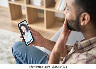 Young Happy Indian Hispanic Handsome Man Holding Cellphone Giving Kiss Sitting On Sofa Dating Online Having Live Videocall With Girlfriend Using Mobile Phone App Technology For Remote Relationships.