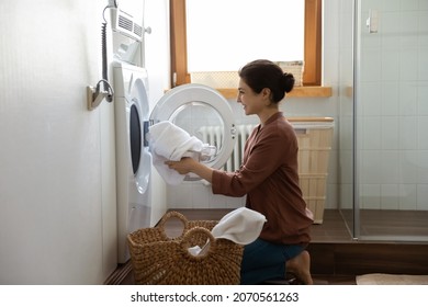 Young happy Indian housewife doing housework putting clothes into washing machine in cozy domestic laundry room. Routine house work, housekeeping, washer-dryer modern appliance or detergent ad concept