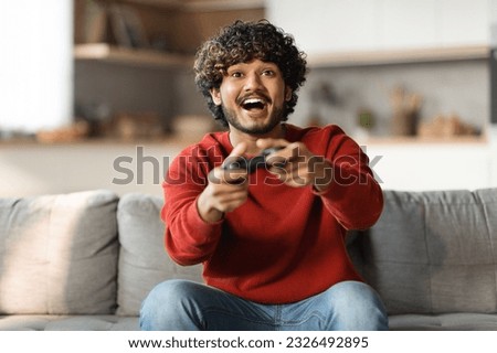 Young happy indian man laughing and playing video games on weekend, sitting on couch alone at home, holding joystick, having fun, copy space. Modern technologies and entertainment concept