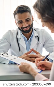 Young Happy Indian Medical Worker Therapist In White Robe Having Appointment Consulting Older Senior Patient Sitting At Desk Filling Data Form In Modern Clinic Hospital. Medical Healthcare Concept.