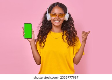 Young happy Indian woman teenager in wireless headphones holding mobile phone and smiling looking at camera demonstrating green screen of gadget stands on pink background. Music playlist concept - Powered by Shutterstock