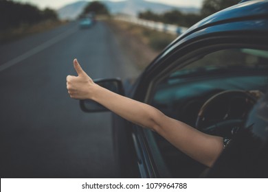 Young Happy Lady Driving A Car And Making Like / Ok Sign With Hand Out Of Window. Girl Driving And Enjoying Road Trip And Travel, Toned With Soft Autumn Colors.