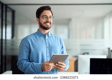 Young happy latin business man leader, company employee, professional businessman manager or software developer holding tab technology looking away standing in office thinking of future work ideas. - Powered by Shutterstock
