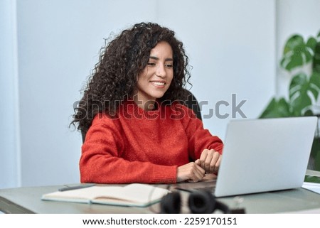 Young happy latin business woman employee using laptop, having remote virtual work meeting call in office or watching webcast online webinar training web course looking at computer sitting at desk.