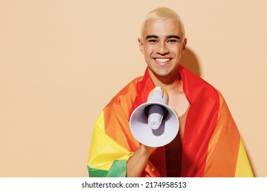Young happy latin gay man with make up in beige tank shirt wrapped in rainbow flag hold shout aside in megaphone isolated on plain light ocher background studio portrait People lgbt lifestyle concept