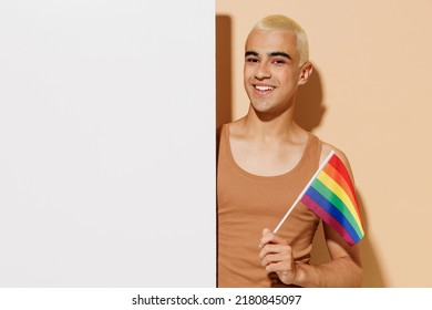 Young Happy Latin Gay Man In Beige Tank Shirt Hold Big White Empty Blank Poster Billboard For Promotional Content, Place For Text Or Image Rainbow Flag Isolated On Plain Light Ocher Background Studio