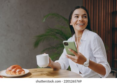 Young happy latin woman 30s wear white shirt hold use mobile cell phone chat hold cup drink tea look aside sit alone at table in coffee shop cafe restaurant indoors. Freelance office business concept - Powered by Shutterstock