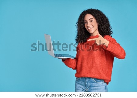 Young happy latin woman pointing at laptop isolated on blue background. Smiling female model holding computer presenting advertising job search or ecommerce shopping website.