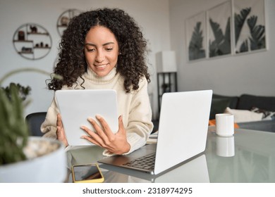 Young happy latin woman, smiling curly casual girl student using digital tablet and laptop elearning or hybrid working at home online looking at tab technology device sitting at table in living room.