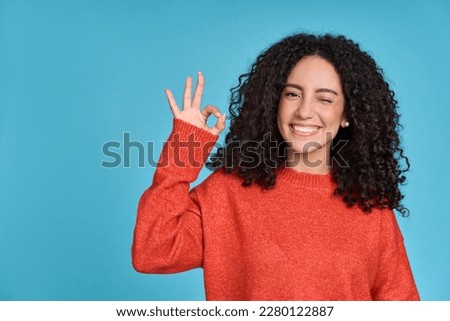 Young happy latin woman student showing ok hand okay sign winking isolated on blue background. Smiling female model advertising sale offer presenting promotion recommending discounts concept.