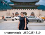 Young happy Latina woman in a black dress and backpack smiling near Gwanghwamun Gate in Seoul, South Korea.