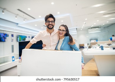 Young Happy Love Couple Standing Hugged With Big Cartoon Box After Buying A New Electronic Product In The Tech Store.